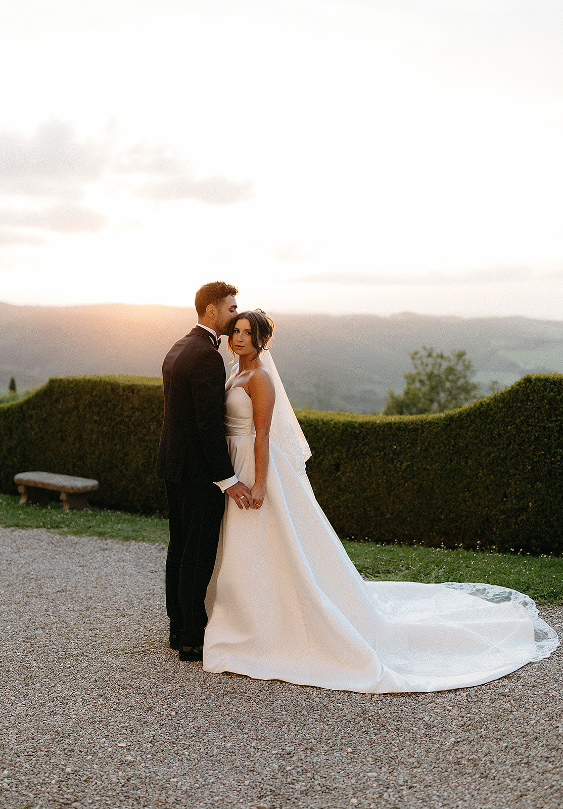 Bride and groom standing at sunset with scenic landscape in the background, captured in a romantic wedding portrait